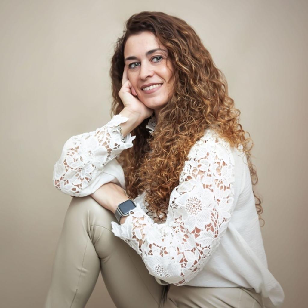 Retrato profesional de una mujer sonriente con cabello rizado largo, apoyada sobre su rodilla, vestida con una blusa blanca de encaje, en un fondo beige.